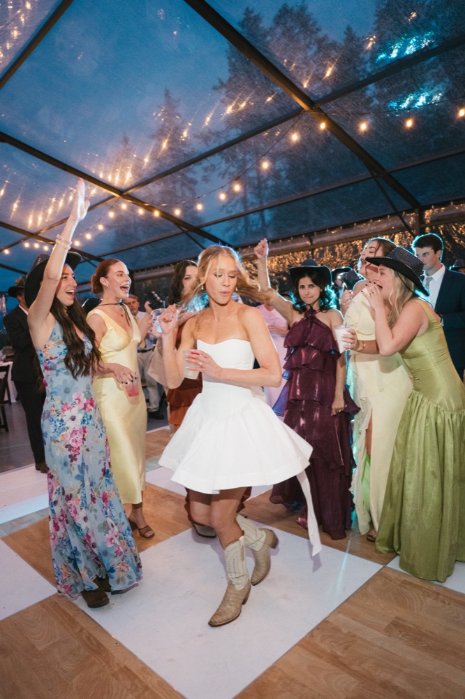 Bride in a short white dress and cowboy boots dancing, friends cheering around her under string lights