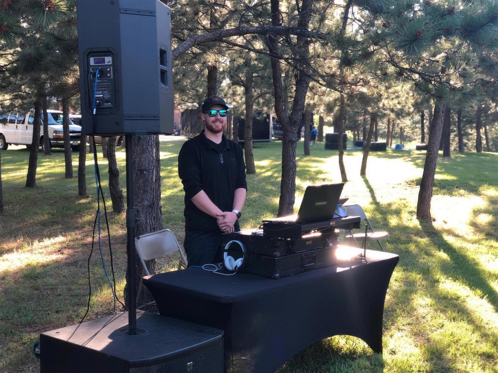 DJ at an outdoor setup with a speaker on a stand among pine trees