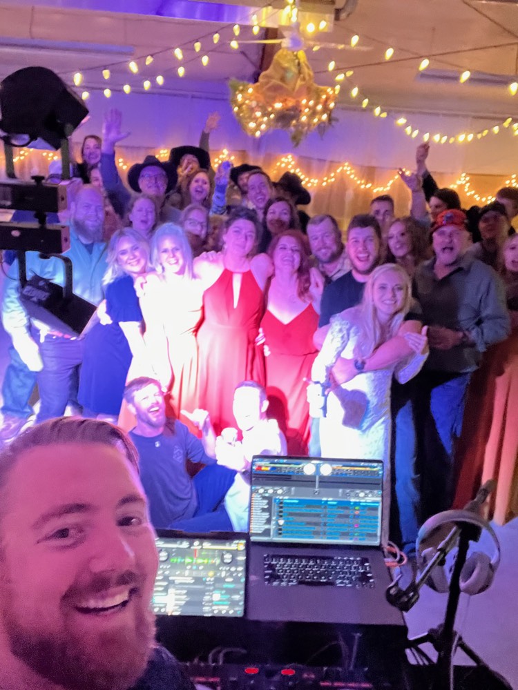 DJ selfie with a packed crowd behind the booth under string lights