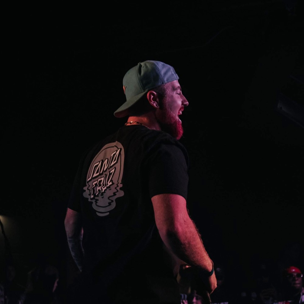 DJ in a cap seen from behind on stage, microphone raised